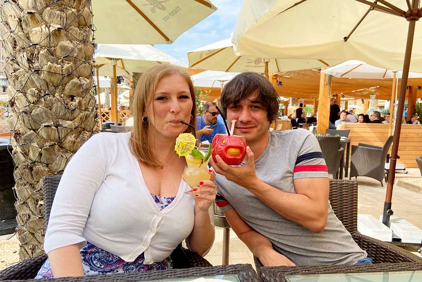 A couple drinking colourful cocktails sat at a restaurant in Dubai with palm trees and umbrellas in the background