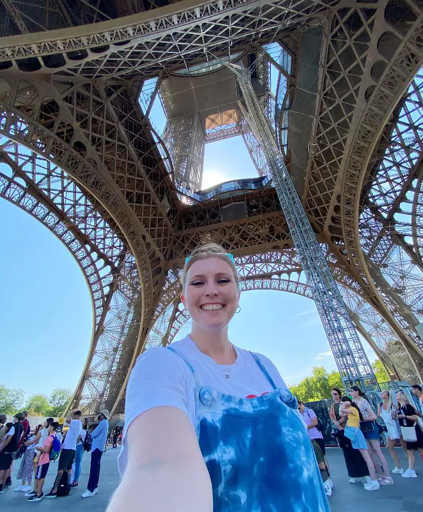Mel taking a selfie wearing blue overalls underneath the bottom of the eiffel tower in Paris with crowds in the background