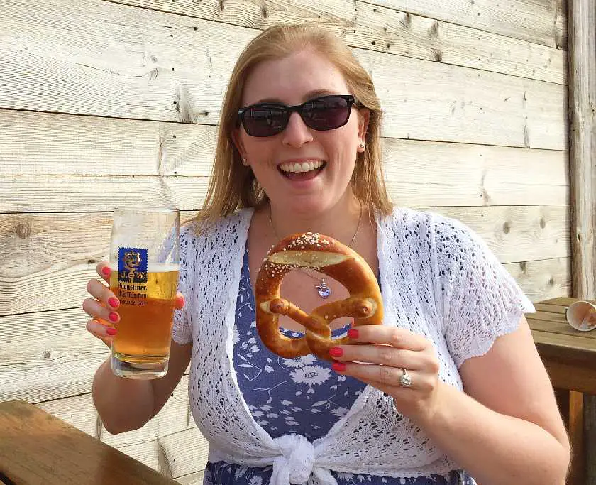 Mel holding up a pretzel and beer sat at a wooden bar in Germany wearing sunglasses and a blue and white dress