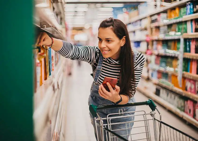 Woman pushing a supermarket trolley picking out food products off a shelf whilst holding her phone