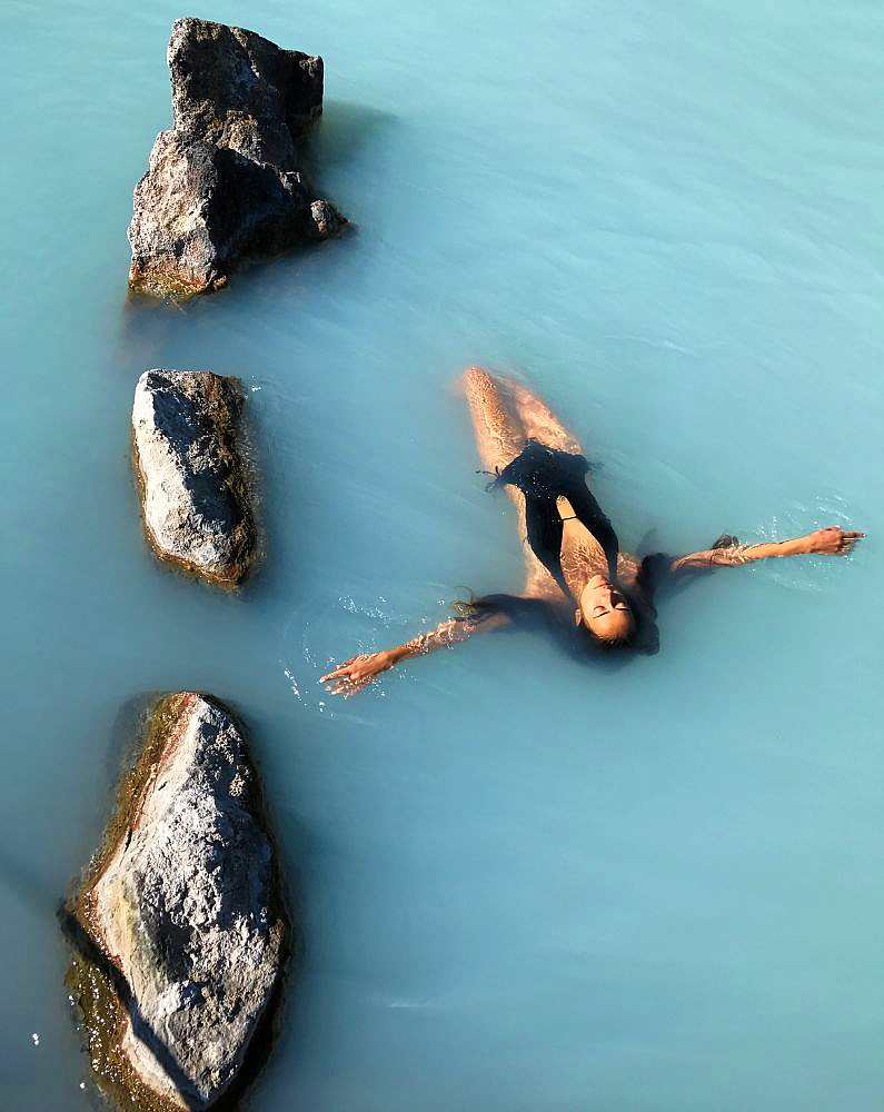 Woman with long dark hair floating on her back relaxing in the bright blue waters of the Blue Lagoon wearing a black swimming costume 