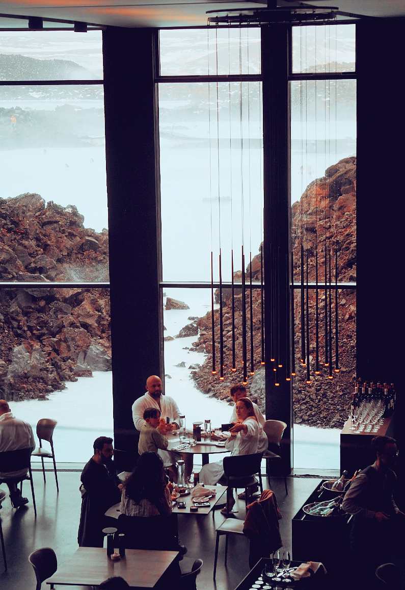 Inside a restaurant in the Blue Lagoon  with people at several tables eating with a view of the Blue Lagoon through the tall, bright windows