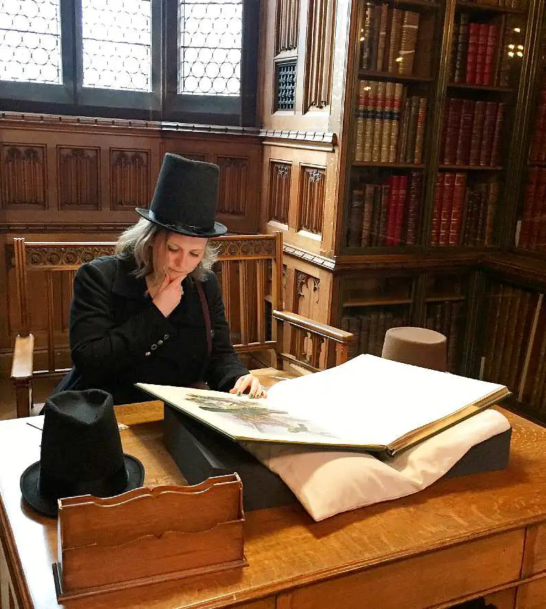 Mel wearing a top hat and pretending to read a large book inside Manchester library 