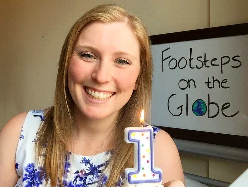 Mel holding a cupcake with a "1" candle with a sign in the background with "Footsteps on the Globe" written on it