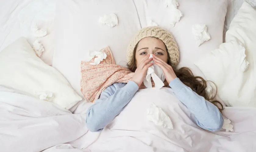 A woman with brown hair wearing a bobble hat and scarf in bed blowing her nose