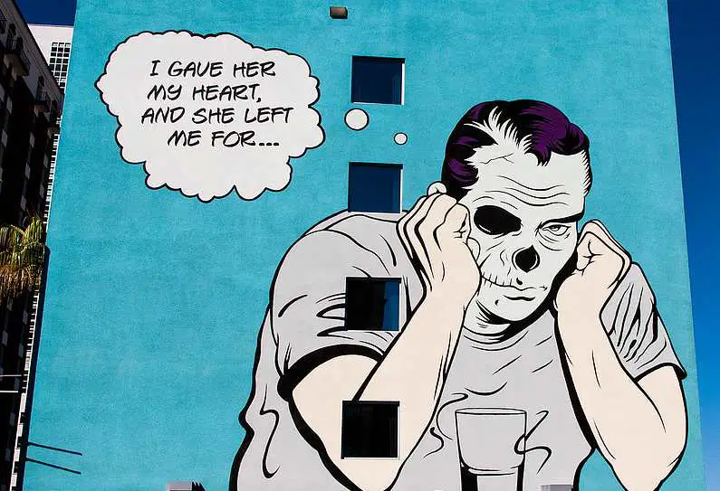 Street art of a man with a skull face holding his hands up to his face against a light blue wall with the thought bubble reading: "I gave her my heart, and she left me for..."