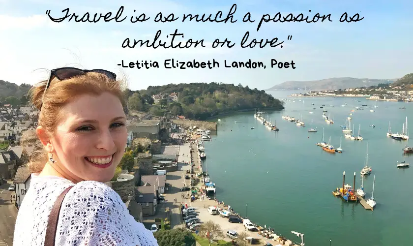 Mel smiling in front of a harbour town in Wales on a sunny day with the quote "Travel is as much a passion as ambition or love." by Letitia Elizabeth Landon an English Poet and Novelist at the top of the image 