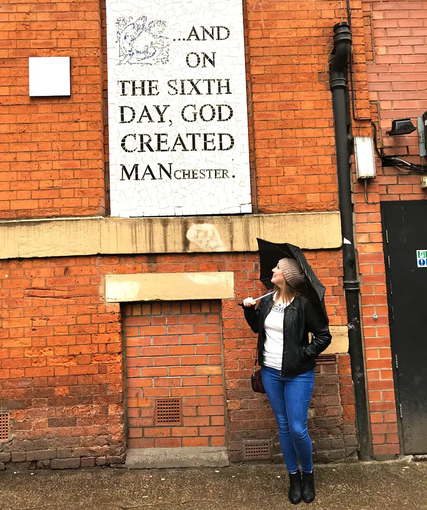 Mel from Footsteps on the Globe, in Manchester holding an umbrella under a sign that says: 'And on the sixth day God created Manchester', Why I took a year off blogging 