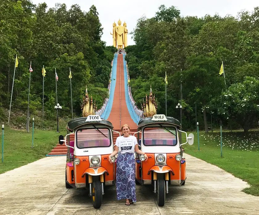 Mel from Footsteps on the Globe posing alongside two orange Tuk Tuk's in front on a long and high staircase leading to three gold buddha statues at the top of the hill in Chiang Mai in Thailand Breaking up, backpacking and beginning again 