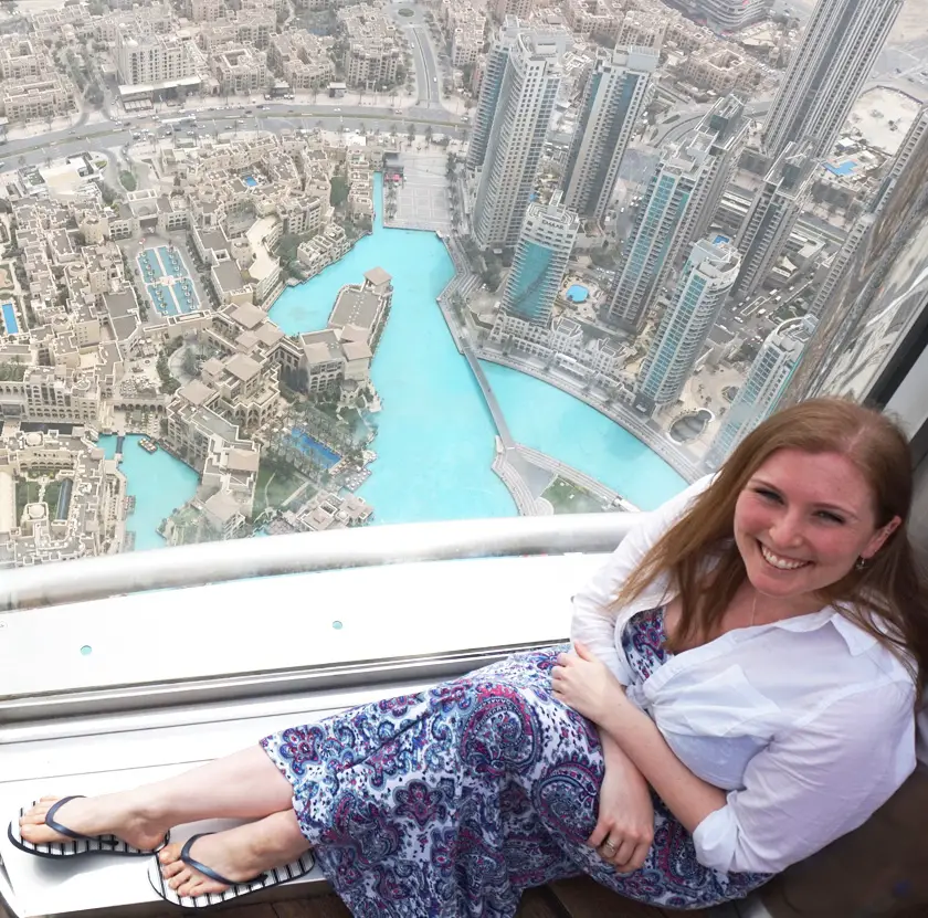 Mel sat on the ground looking up at the camera with a view of Dubai's busy city in the foreground from the top of the Burj Khalifa.
