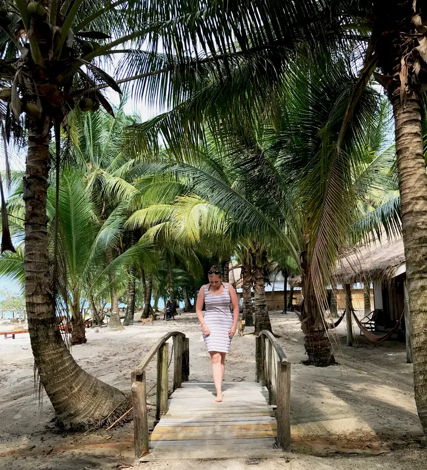 Mel walking over a bridge on an sandy island with palm trees in the background