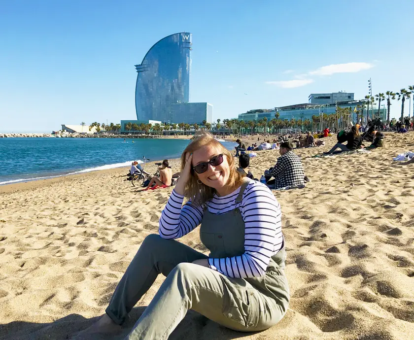 Mel from Footsteps on the Globe sat on Barcelonetta Beach in Barcelona Spain in front of the W Hotel in green dungarees and sunglasses, Breaking up, backpacking and beginning again