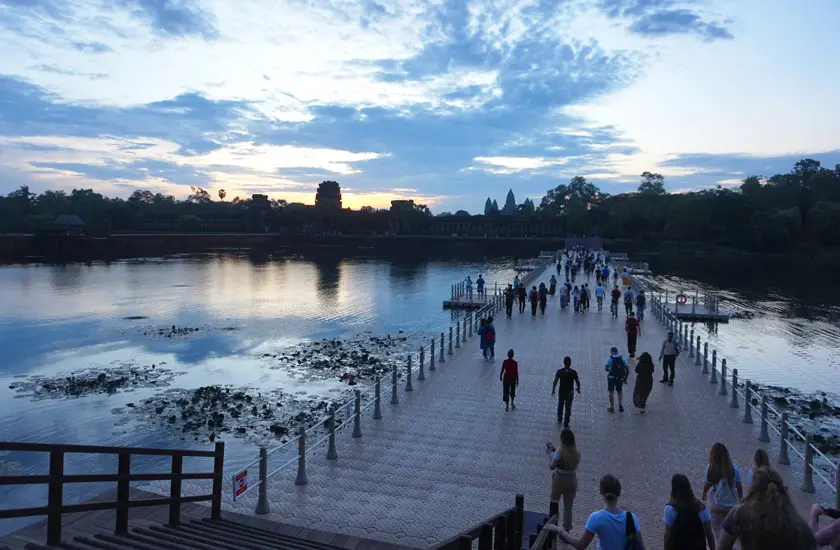 Walk way to Angkor Wat across the moat at sunrise with blue skies and people walking across. 