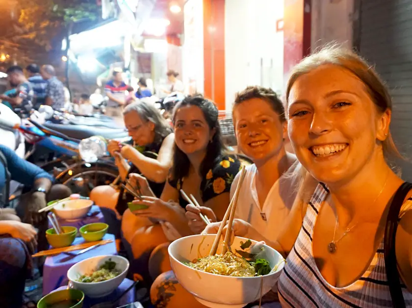 Girls having street food sat on the street holding bowls of noodles