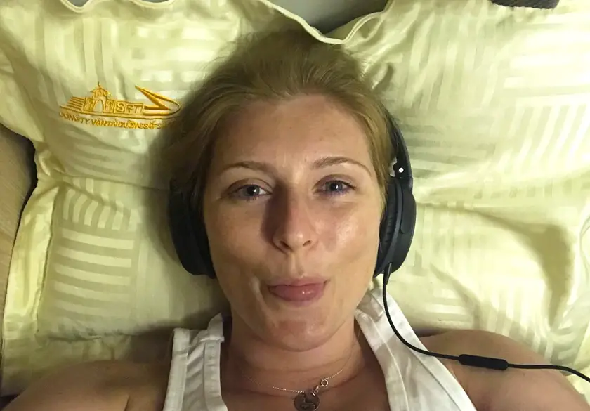 Mel from Footsteps on the Globe lying on the bottom bunk on the overnight train in Vietnam with headphones on resting on a yellow pillow and sticking her tongue out taking a selfie