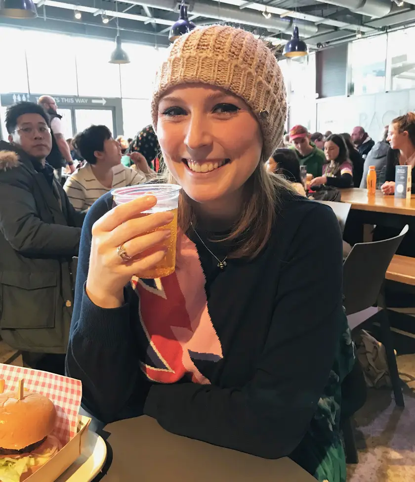 Mel holding a vegan butterbeer wearing a pink wooly hat and navy jumper in The Backlot Cafe 