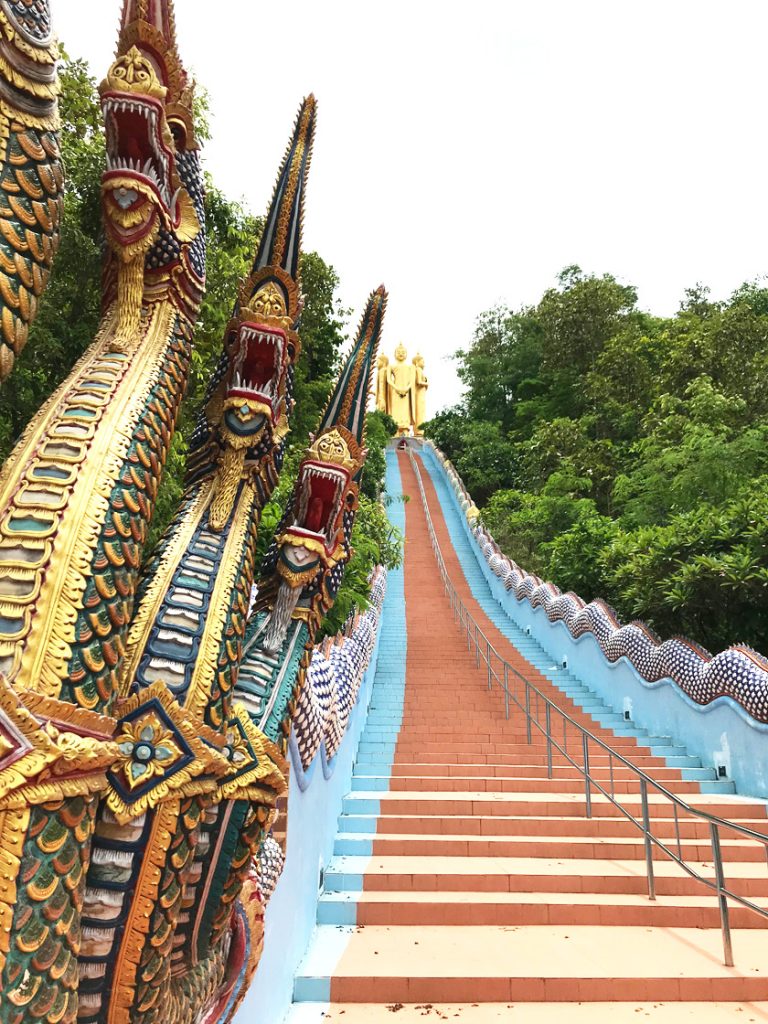 Golden dragons next to a red and blue staircase leading to three golden buddha statues at the top of a hill