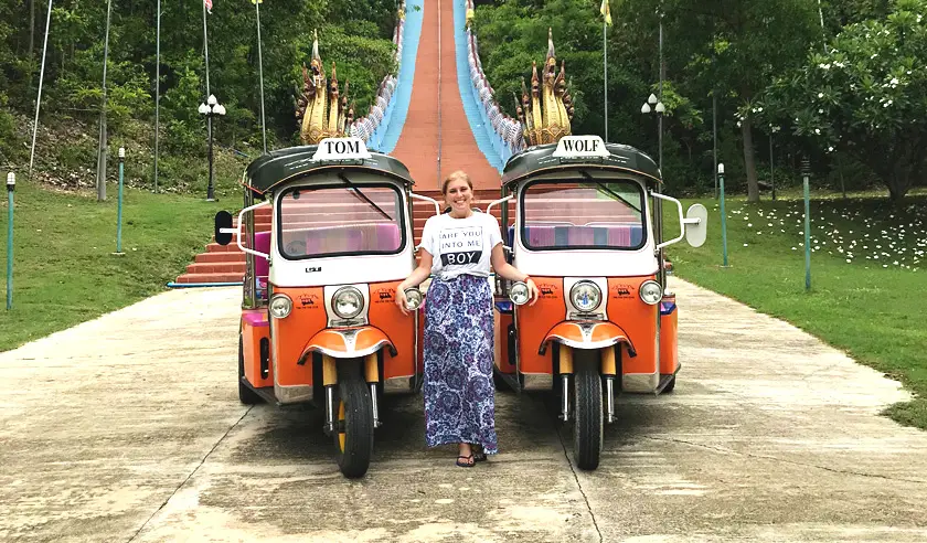 Mel from Footsteps on the Globe standing betweentwo orange and white tuk tuks in front of a temple's red and blue stairs in Chiang Mai Thailand
