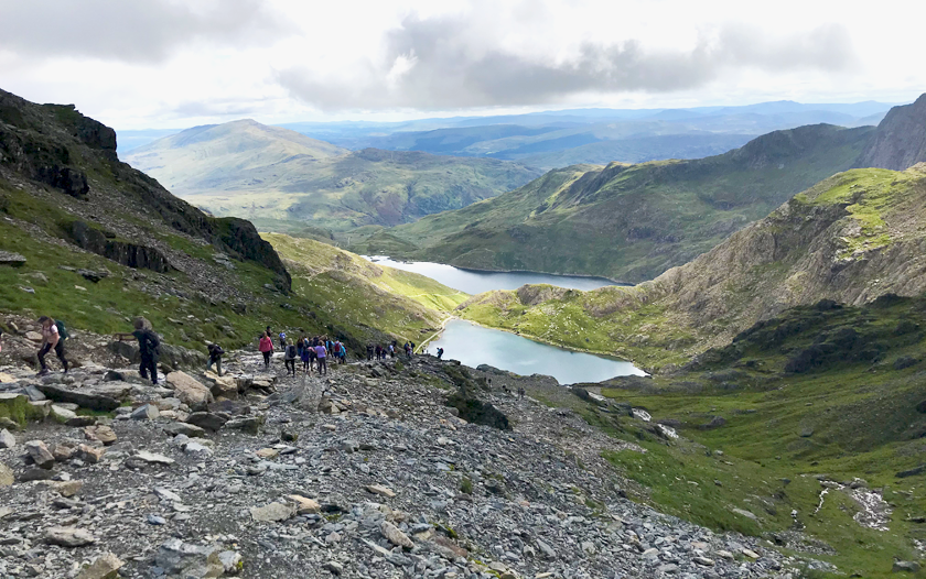 Looking down over the Miners Track where a line of people in the distance on a rocky trail with lakes and other Snowdon mountain peaks in the background