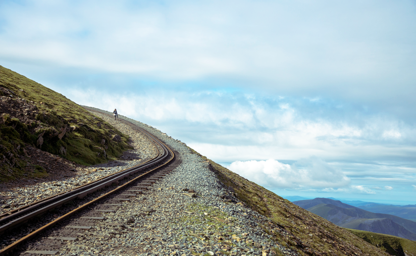 Railway tracks at the top of Snowdon with a lone climber and clouds in the background.