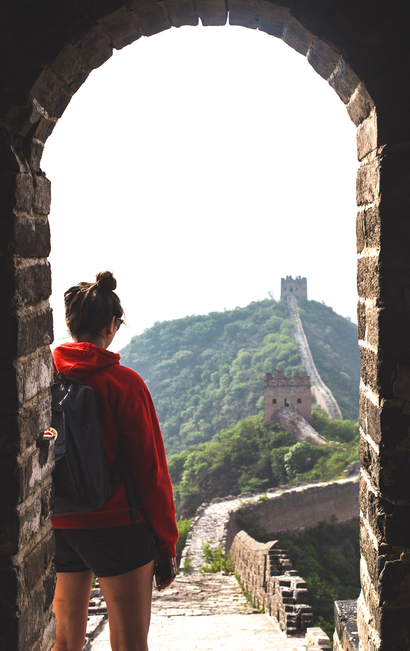 The Great Wall of China from the point of view of an archway with a woman wearing a red hoodie and black backpack in the foreground
