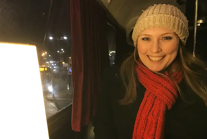 Mel sat on a dark bus at night smiling next to a glowing light wearing a red scarf and wooly hat