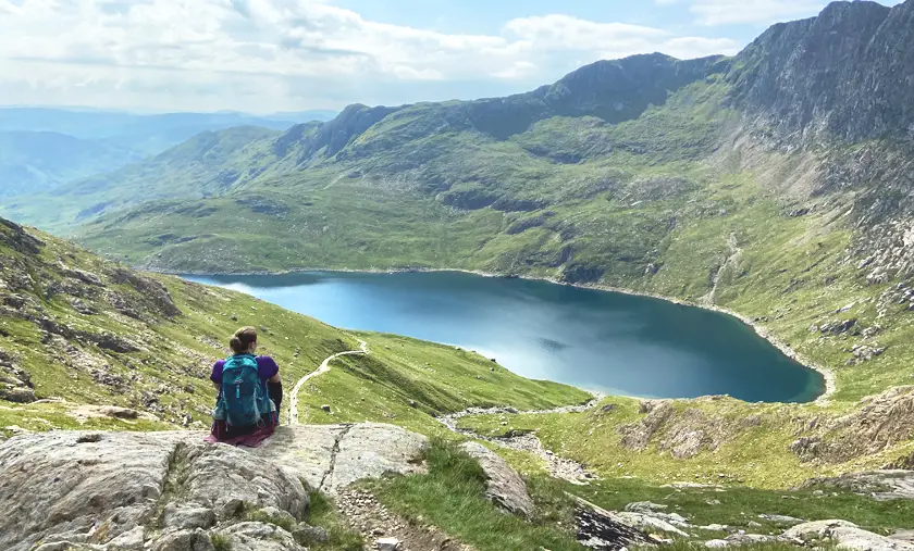 Mel with her back to the camera, sat looking out over a beautiful blue lake on the Pyg Track at Snowdon