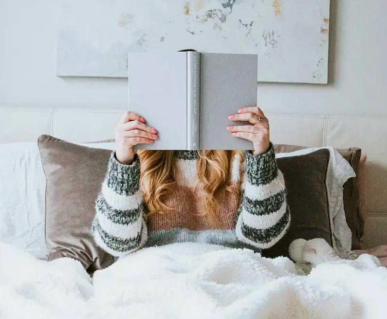 Woman wearing a stripped woolly jumper, sat in her bed with a grey book held up over her face