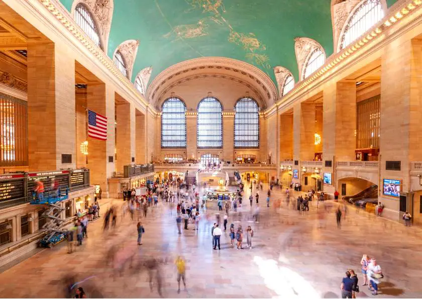 Inside Grand Central Station in New York with people walking around the main concourse, high arched windows and turquoise ceiling