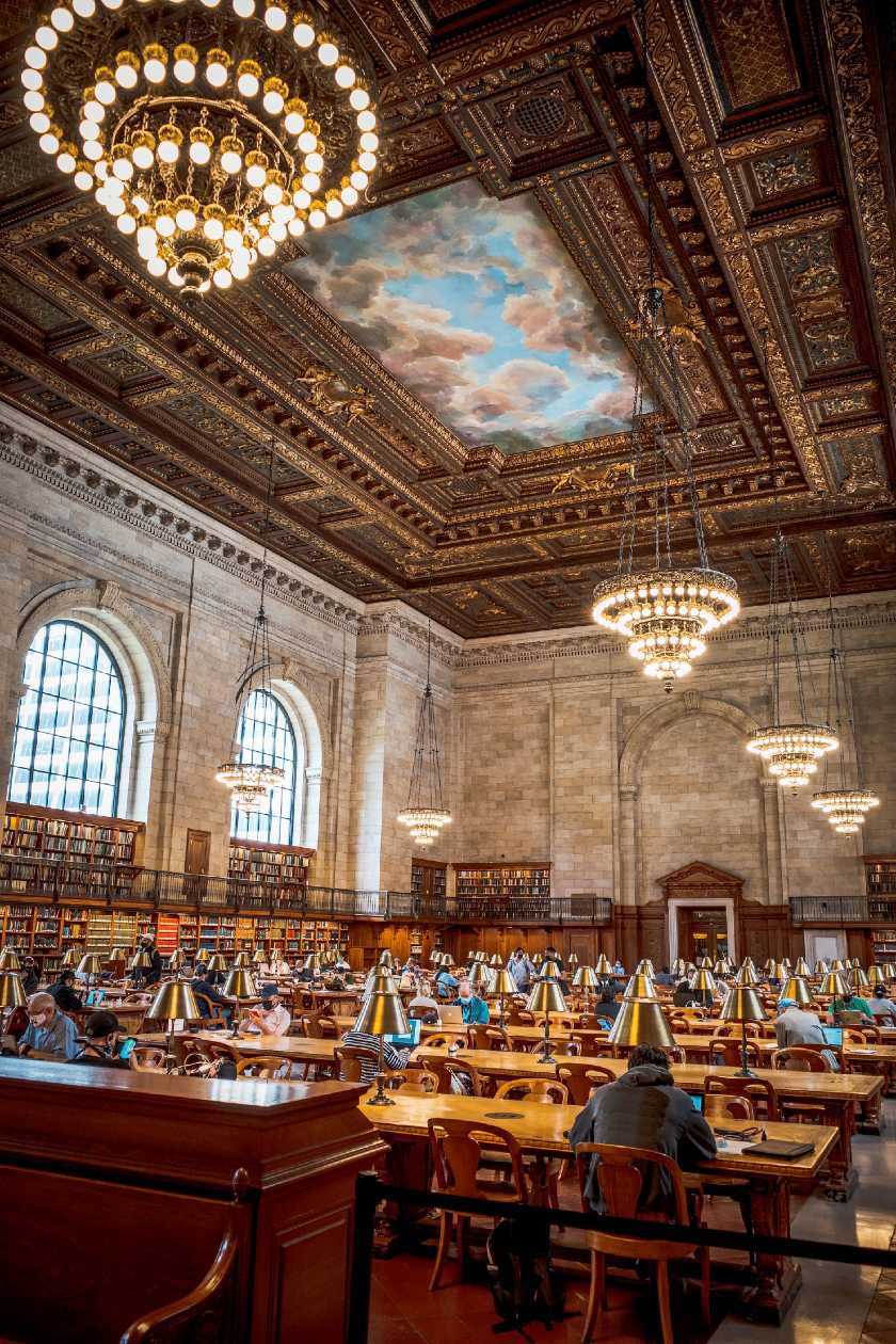 Inside the New York Public library with rows of people studying at desks, hanging lights and and a painted ceiling mural