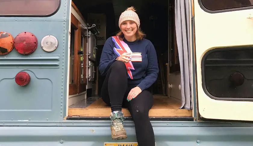 Mel sat at the back of an open door blue converted school bus holding a cup of tea wearing a wooly hat and hiking boots