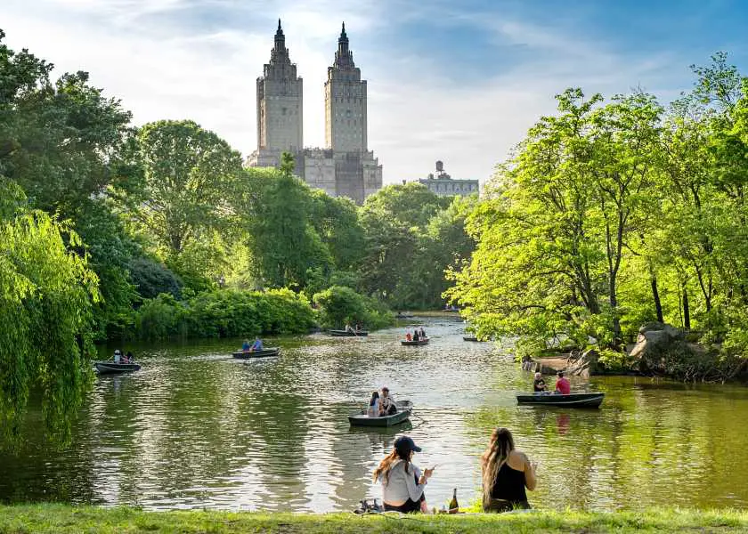 People sat by Central Park lake in New York with The Plaza hotel in the background