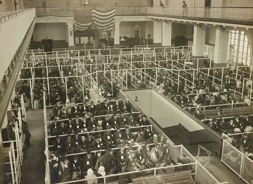 Old black and white photo of iside the processing building on Ellis Island with people queuing sat down to have their paperwork done