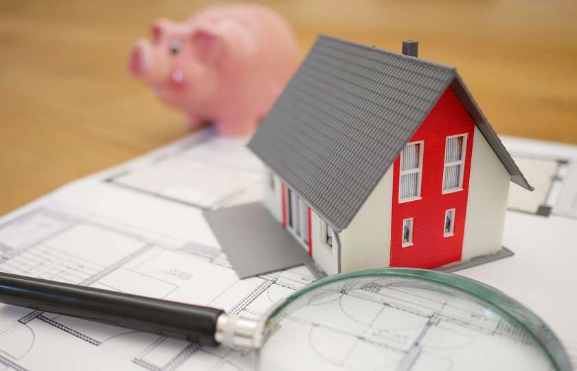 Model of a house with piggy bank in the background and magnifying glass in the foreground on top of architectural drawings
