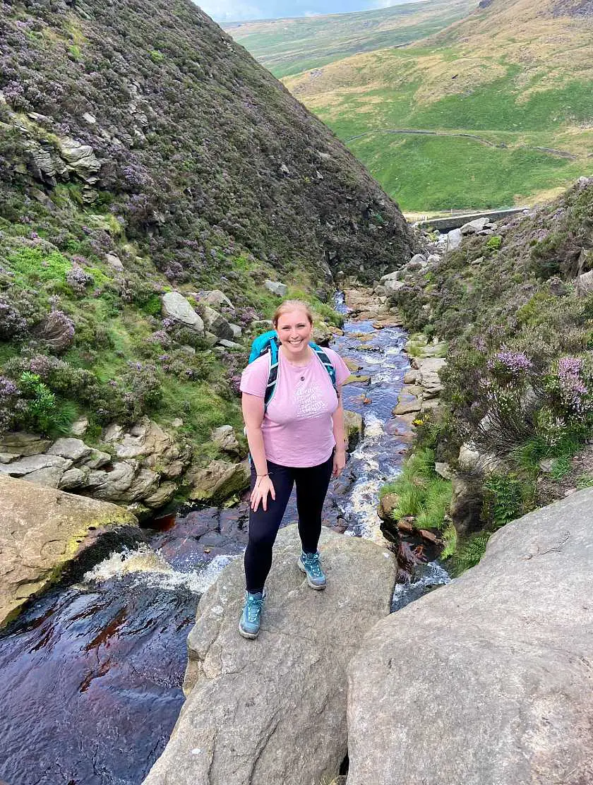 Mel wearing a pink tshirt, green hiking boots and backpack, hiking over a stream in Dovestones Reservoir between two green peaks