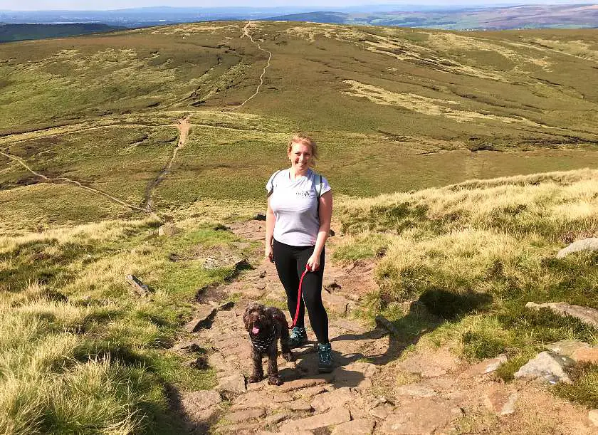 Mel stood on a walking trail in the peak district with the green hills behind her holding Dexter the dog by a lead