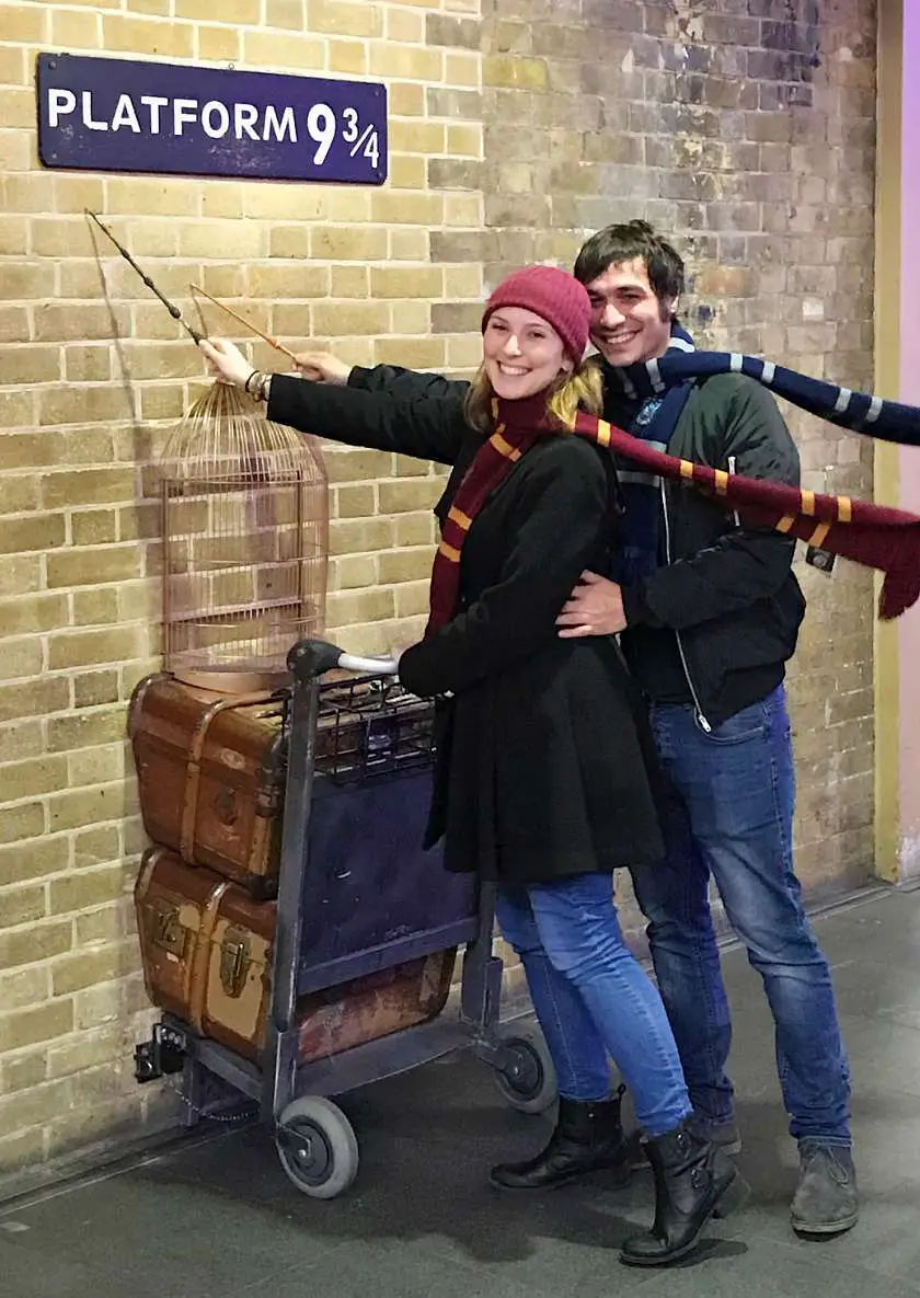 Mel and Joe holding wands up to Platform 9 and three quarters at King's Cross Station wearing Hogwarts scarves and pushing a trolley with luggage