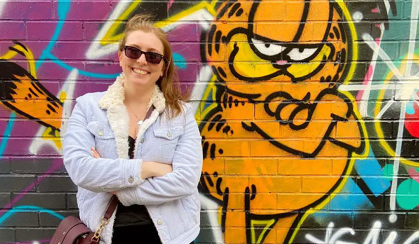 Mel crossing her arms against a wall of street art featuring Garfield the cat
