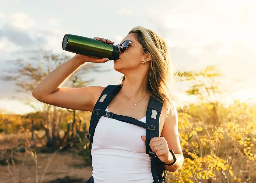Girl drinking from a reuseable water bottle whilst on a hike wearing a white t-shirt with a black backpack and sunglasses