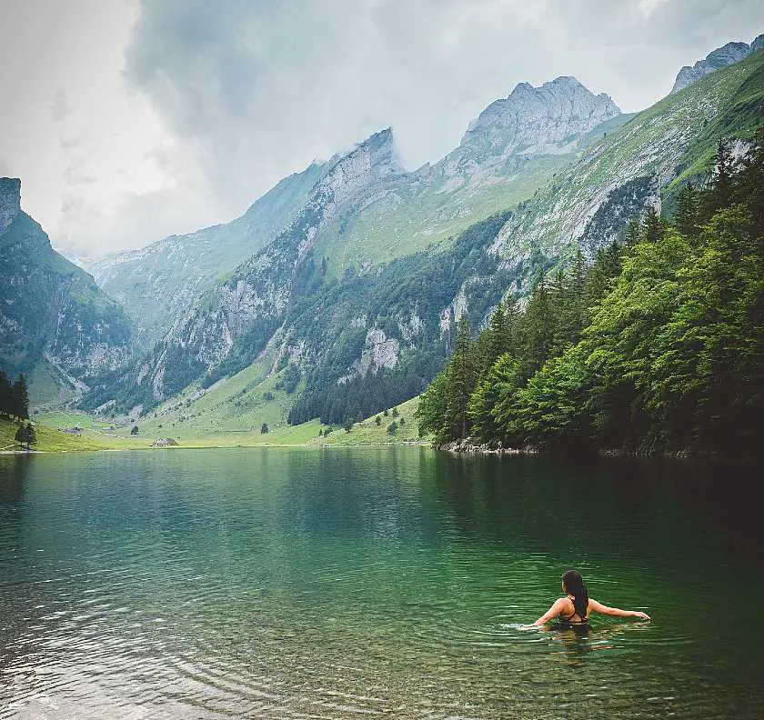 Dark haired girl with her back to the camera wild swimming in a lake with green mountains in the background