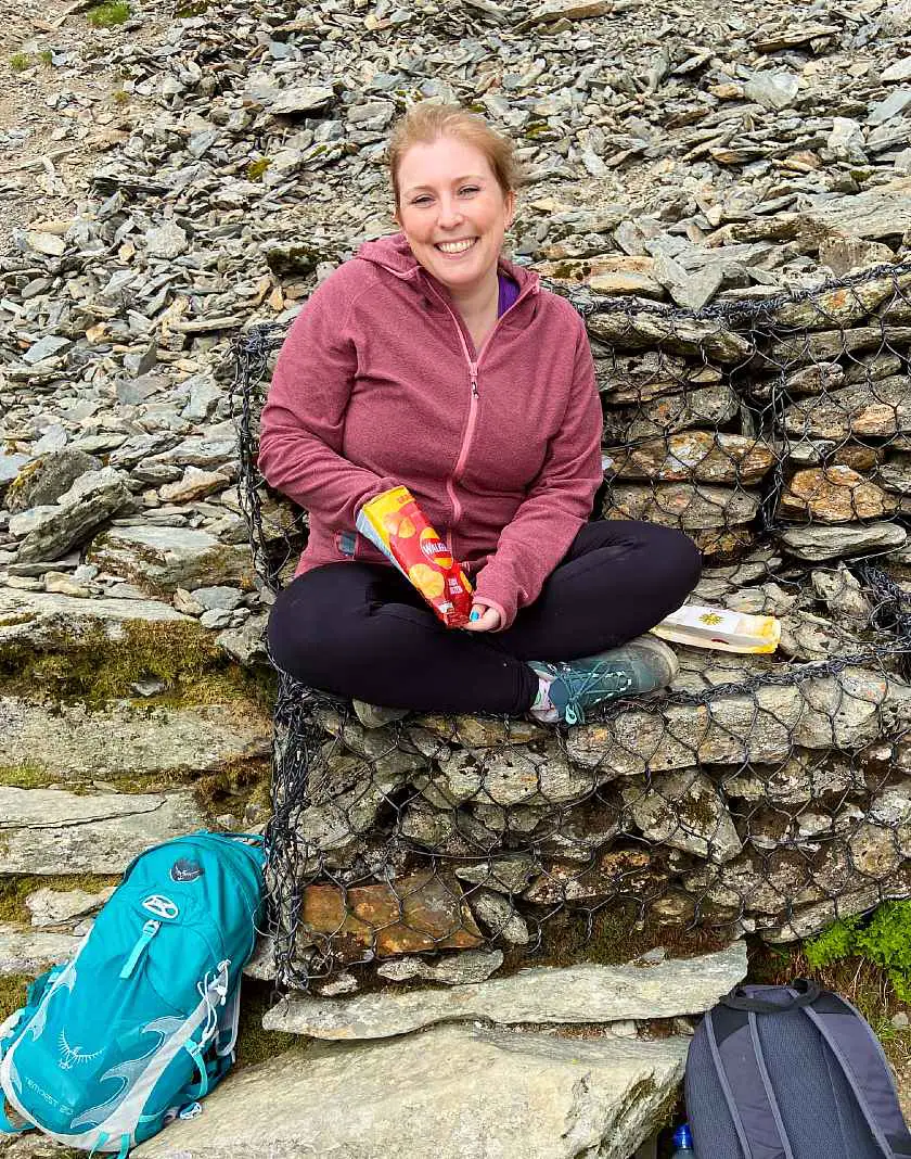Mel sat on a pile of rocks eating crisps before the final ascent of Snowdon with backpacks at her feet
