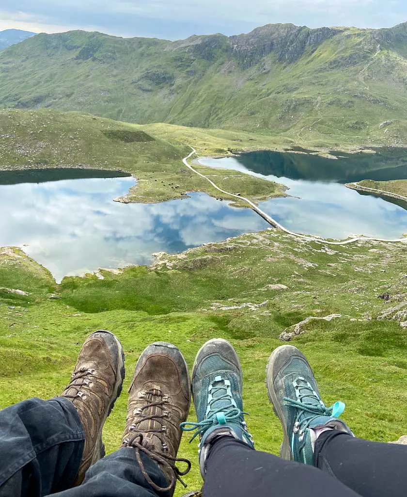 Two people sat down with their hiking boots on front of them with the Pyg Trail's lake and peaks in the background