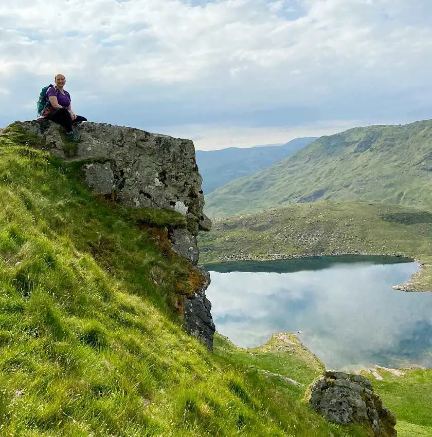 Mel sat on a high rock and grass mound with a lake behind her 