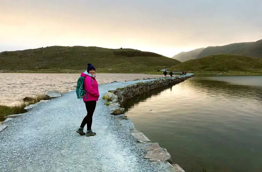 Mel descending via the Miners Track at sunrise, walking over a stone trail across a lake at the bottom of Snowdon wearing a pink jacket and blue bobble hat