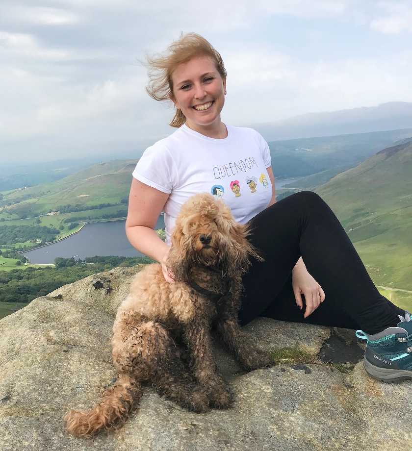Hiker sat smiling at the top of Dovestones Reservoir with her blonde  muddy cockapoo 