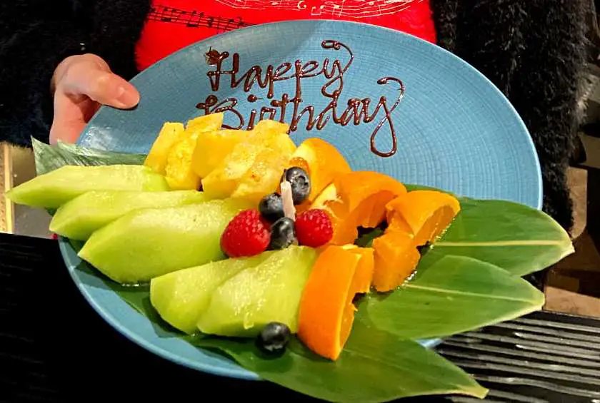 Tropical fruit on a blue plate with a candle in it and "Happy Birthday" written in chocolate sauce