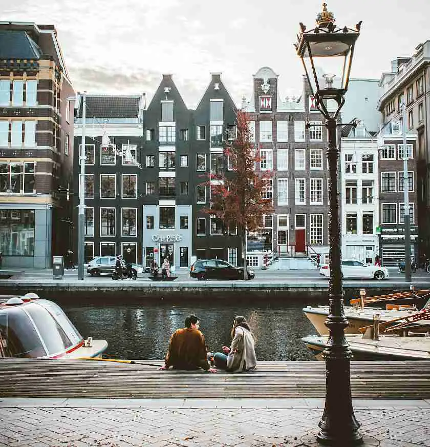 Two people sat by the canal in Amsterdam in front of an old fashioned lantern and row of town houses