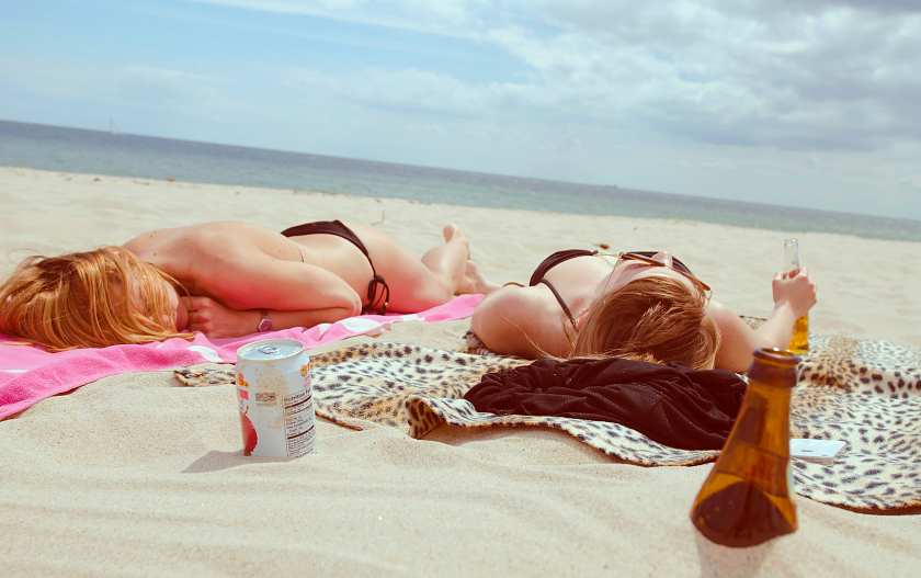 two girls sunbathing on the beach facing the sea lying on colourful beach towels with beer bottles in the sand