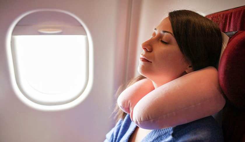 Woman lying back with her eyes closed in her plane seat by a window wearing a travel pillow around her neck