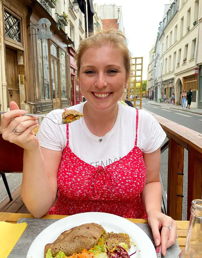 Mel smiling holding up a fork of food sat outside Le Potager du Marais wearing a red dress and a white t-shirt